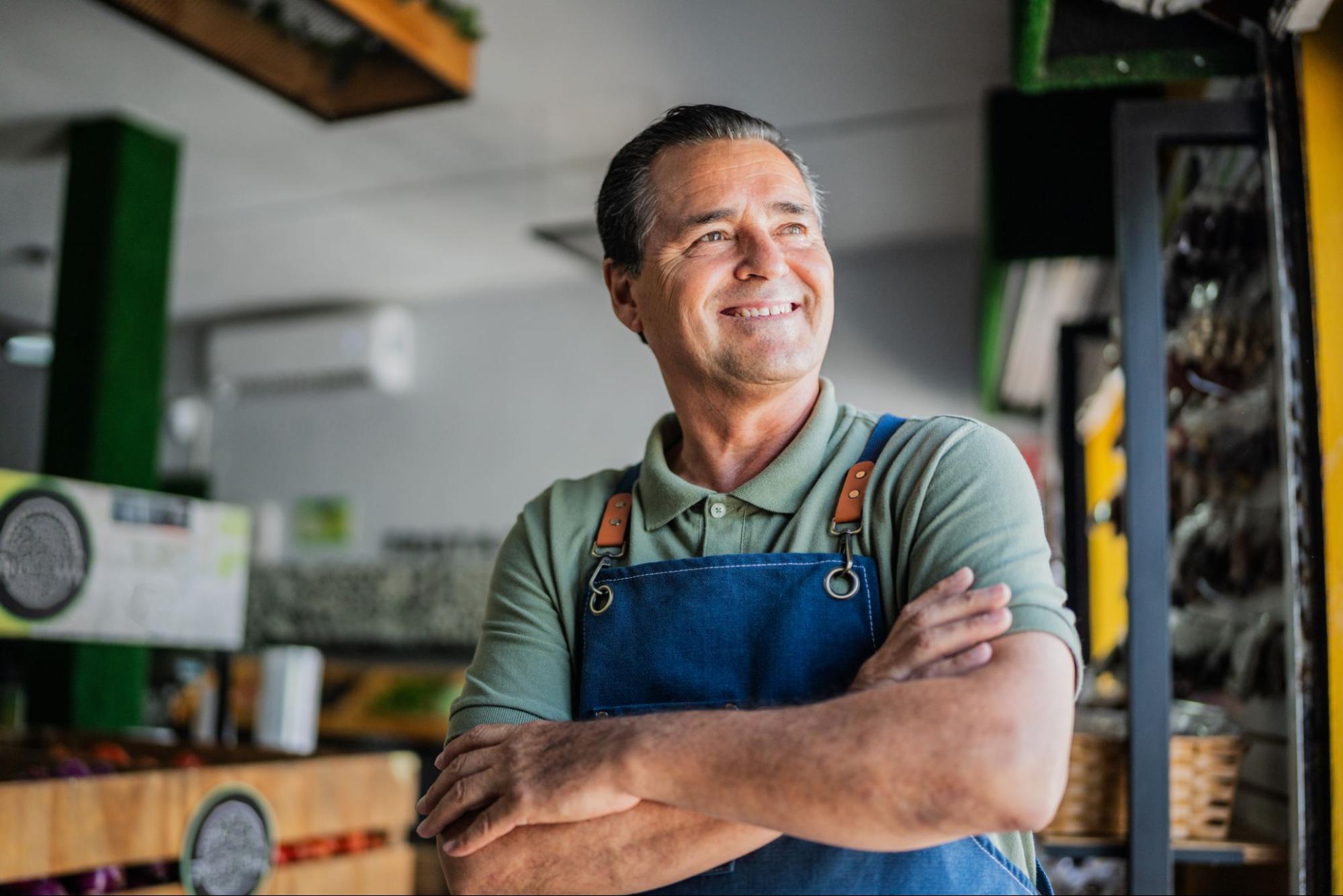 A smiling man wearing a denim apron stands in a workshop with his arms crossed.